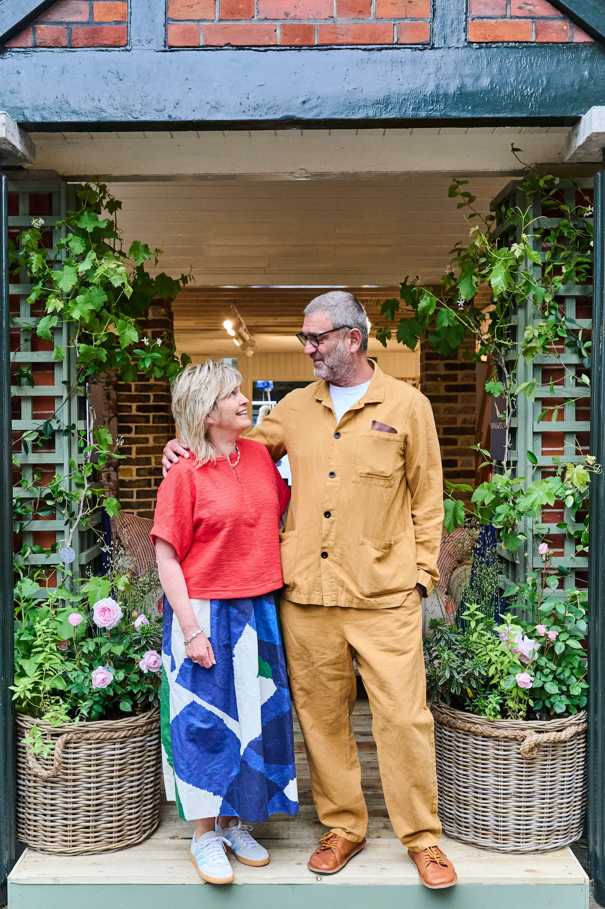 An older couple stands on a porch surrounded by potted plants and flowers. They smile at each other warmly.